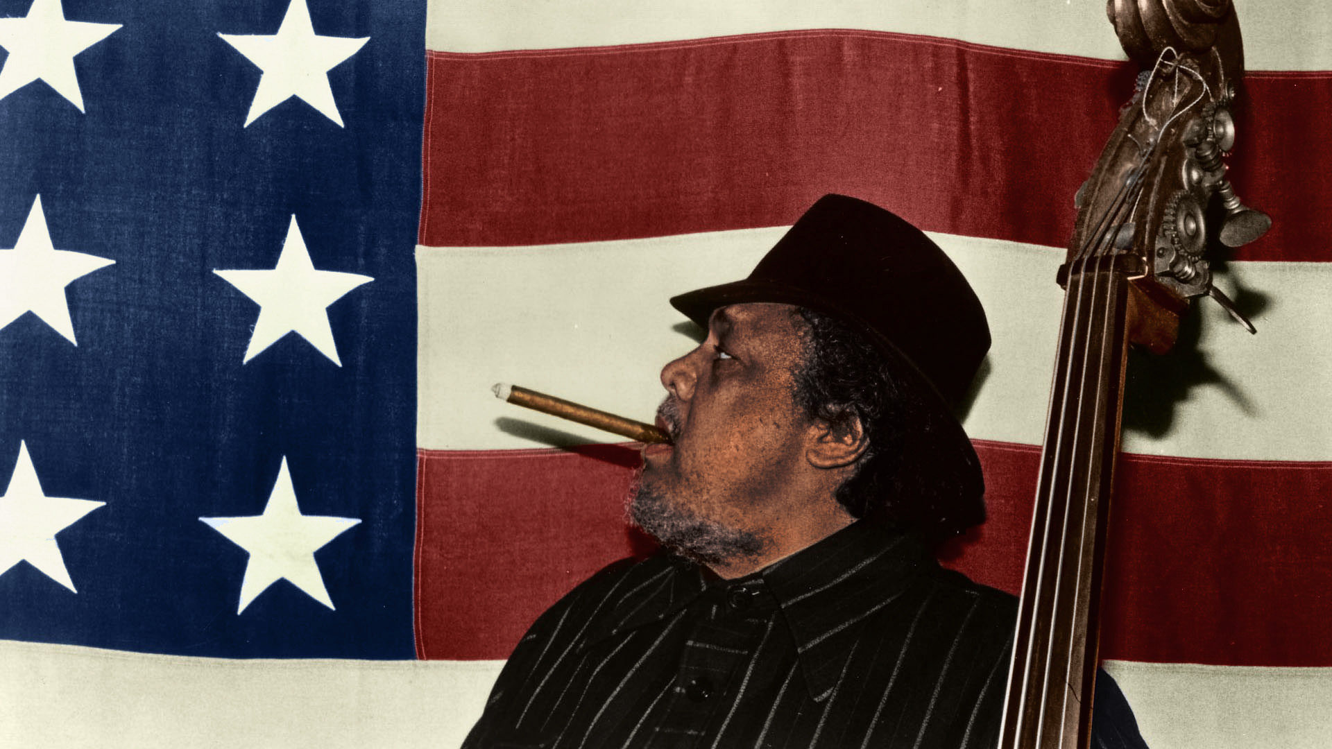 Charles Mingus smoking a cigar in front of the American flag. (Photographed in 1977 by his wife Sue Mingus)